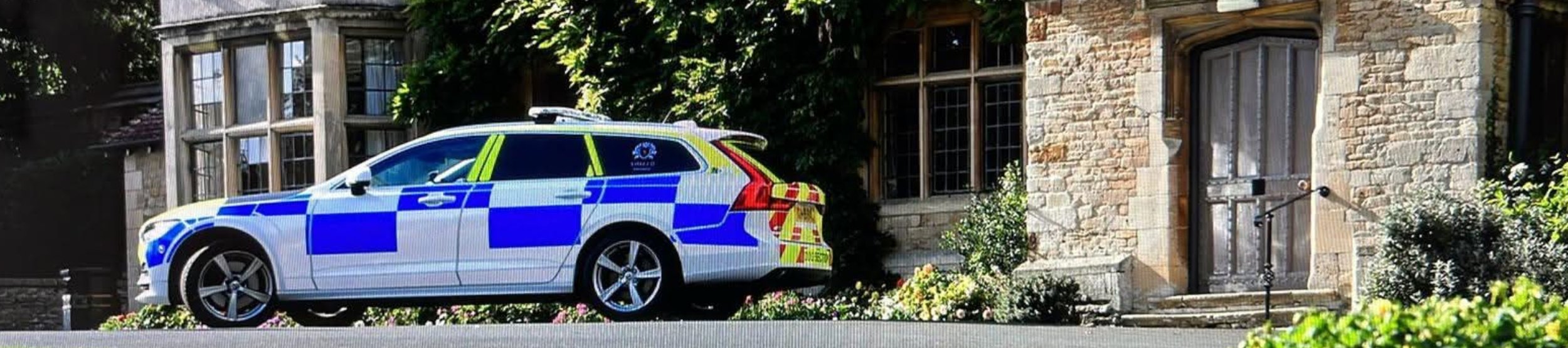 Police car parked in front of a historic stone building with ivy, surrounded by lush greenery and colorful flowers, under clear blue skies.