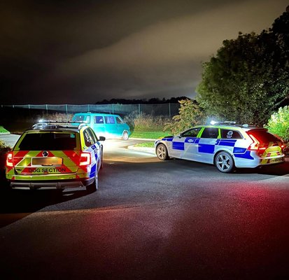 Two police cars with 'Dog Section' markings block a road at night, facing a teal van. The scene is illuminated by their headlights under a cloudy sky.